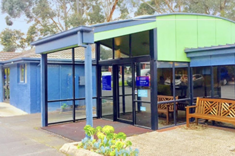 The entrance to Wattletree Specialist Consulting Suites: a glazed porch under a curved lime-green roof, set into a blue rendered single-storey building, with timber benches beside the automatic doors and a level path from the car park.